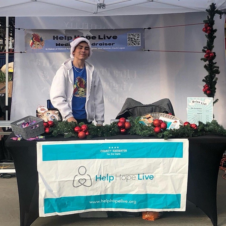 Kevin Lopez's son is standing behind a fundraiser booth that includes a large white and teal Help Hope Live banner with the website www.helphopelive.org and text Charity Navigator Four Star Charity. On the table in front of him is a black tablecloth and a fake pine wreath with red Christmas tree decoration baubles plus some candy, brochures, and a Price List. Kevin's son has light brown skin, dark eyes, a Santa hat, a gray hoodie, and a blue t-shirt that includes a logo that is a stylized hand holding a heart with the Texas and American flags and the text Heart of a Warrior Team Lopez. The same logo appears on a large hanging banner alongside a QR code. The banner says Help Hope Live Medical Fundraiser In Honor of Retired Police Officer and Veteran Kevin Lopez.