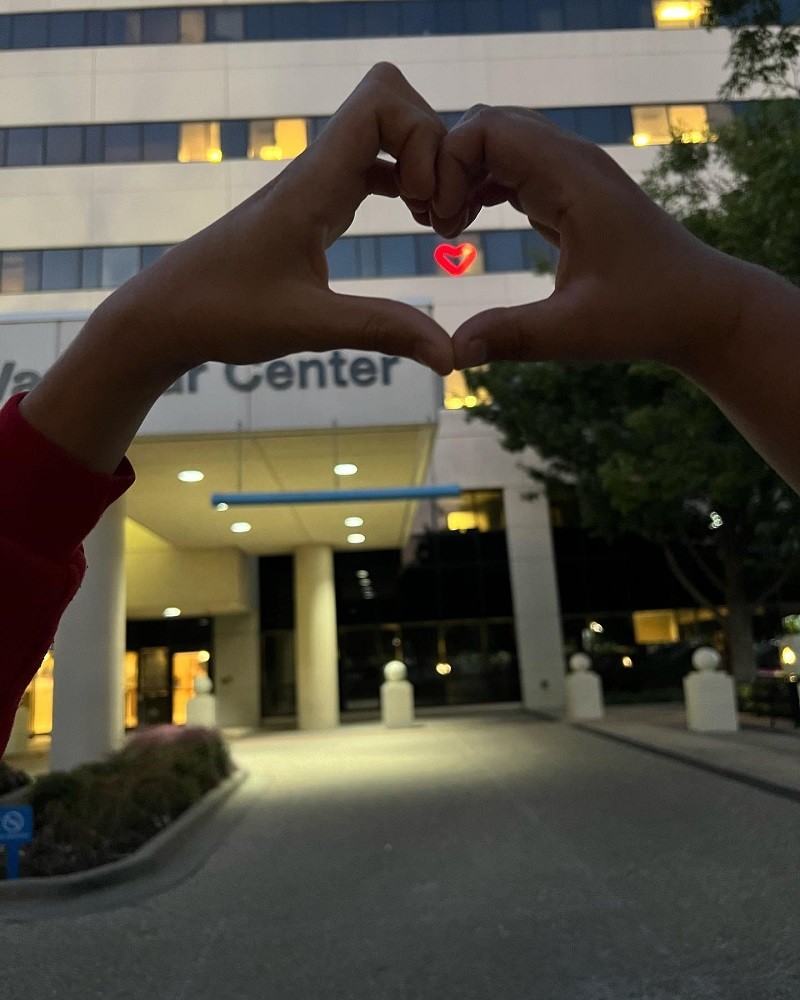 Two light brown hands come together to form a heart - beneath the hand heart is a red lit-up heart in the window of a hospital building. It appears to be dusk.