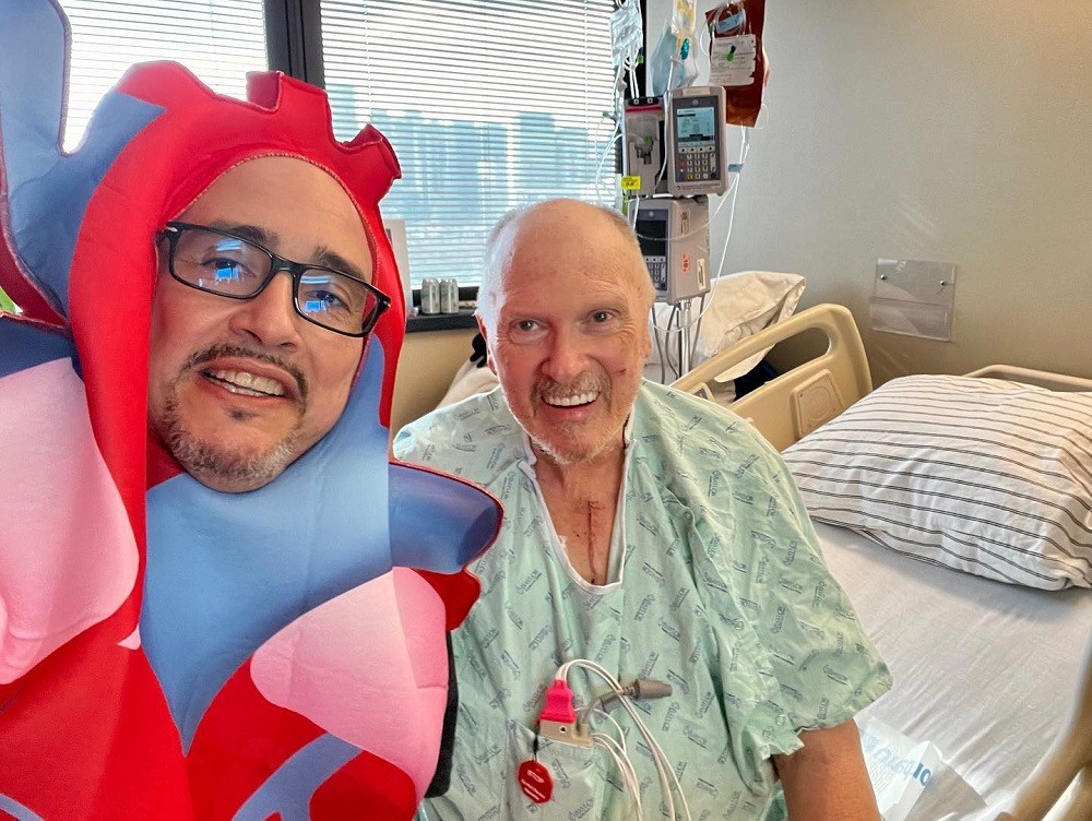 Kevin Lopez is with a man in a hospital room taking a selfie as Kevin wears a goofy anatomical heart costume in blue, pink, and red. He has light brown skin, a salt and pepper goatee, dark eyes, and black-rimmed glasses. The man he is with has light skin, a brown and gray mustache and short beard, blue eyes, and a visible surgery scar extending to the neck of his hospital gown vertically from just under his neck. His hospital gown is light green.