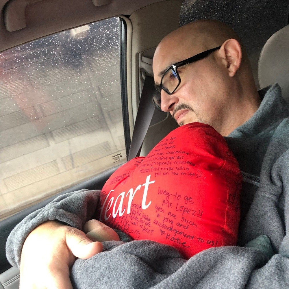 In a photo taken in profile as he sits in the passenger seat of a car, Kevin Lopez hugs a red heart plushie signed by medical staff members and has an expression of discomfort or melancholy on his face. Kevin has light brown skin, dark eyes, a short dark goatee, a bald head, and black rimmed glasses. There is rain on the car windows.