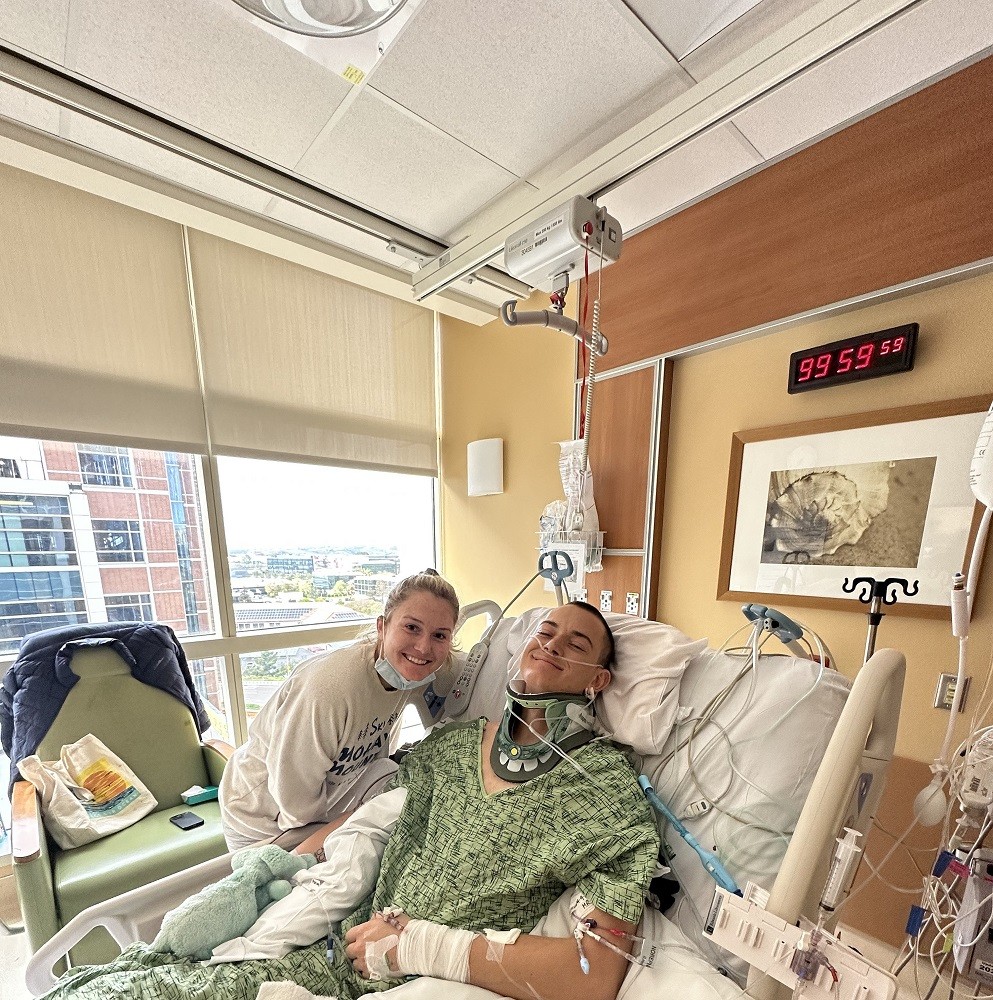 Collin Bosse is in a hospital bed wearing a green hospital gown after a spinal cord injury with a neck brace and medical devices hooked up to him. He is smiling with light skin and short cropped dark hair. His sister is beside him with light skin, a face mask pulled down to her chin, and blonde hair pulled back into a ponytail. A building and city scene is visible from their window (they are not on the ground floor).