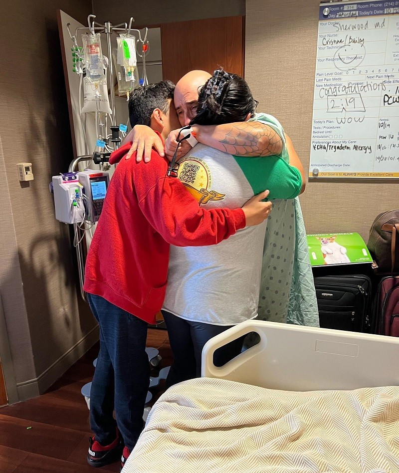 In an emotionally charged captured moment, Kevin Lopez wears a hospital gown and hugs his son and daughter at the same time with an intense expression on his face of fear, love, and resilience. He has light brown skin and a bald head. His son and daughter have light brown hair and wear casual clothes.