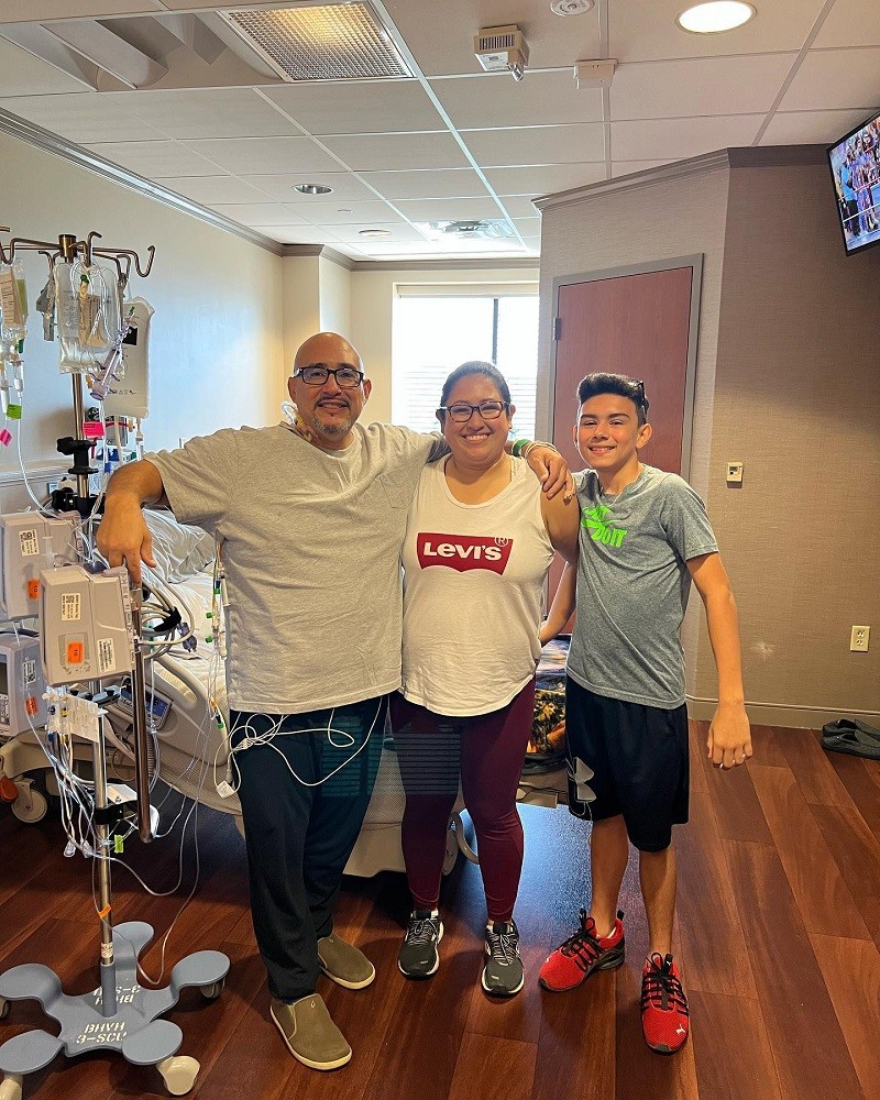 Kevin Lopez and his two children are in a hospital room together while Kevin is hooked up to a medical cart. Kevin has light brown skin, a bald head, a salt-and-pepper goatee, black rimmed glasses, a gray t-shirt, dark sweatpants, and slip-on camo green shoes. Kevin's daughter has light brown skin, black hair pulled back, dark eyes, tortoiseshell glasses, a t-shirt, maroon athletic leggings, and sneakers. His son has light brown skin, short dark hair, dark eyes, a gray t-shirt, athletic shorts, and red sneakers.