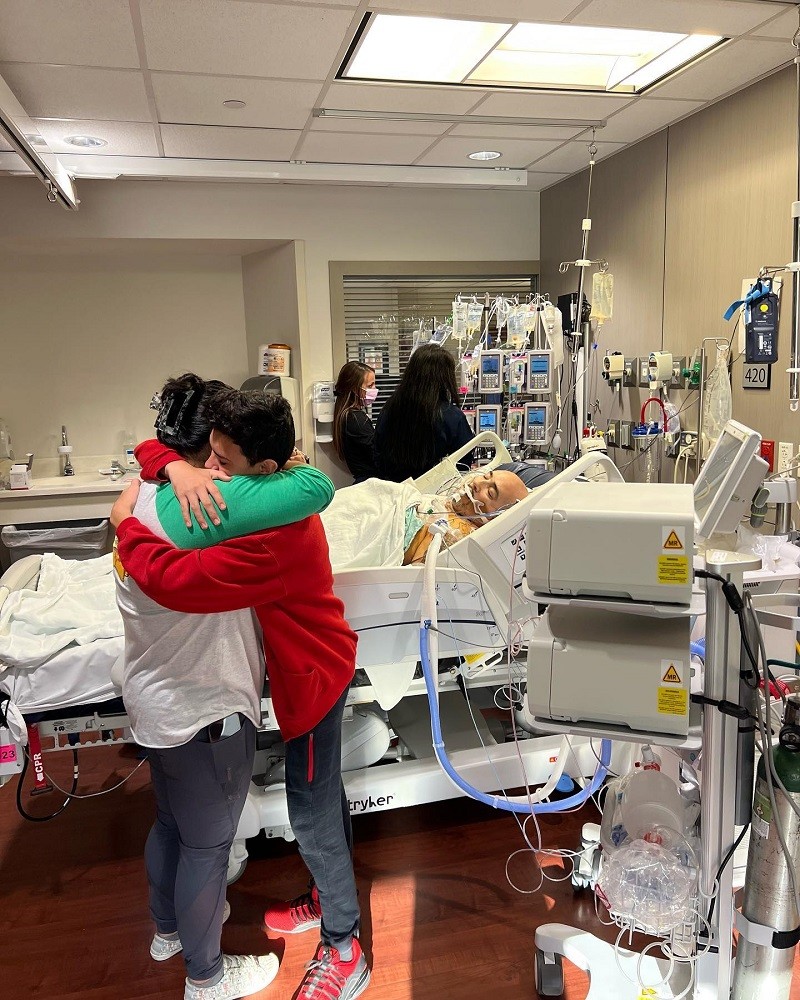 In a hospital room full of equipment and carts, Kevin Lopez lies with his eyes closed and intubated in a hospital bed as his two children hug each other tightly in an emotional moment. Kevin has light brown skin and a bald head. His daughter and son both have light brown skin and dark hair and wear casual athletic clothing as they embrace.