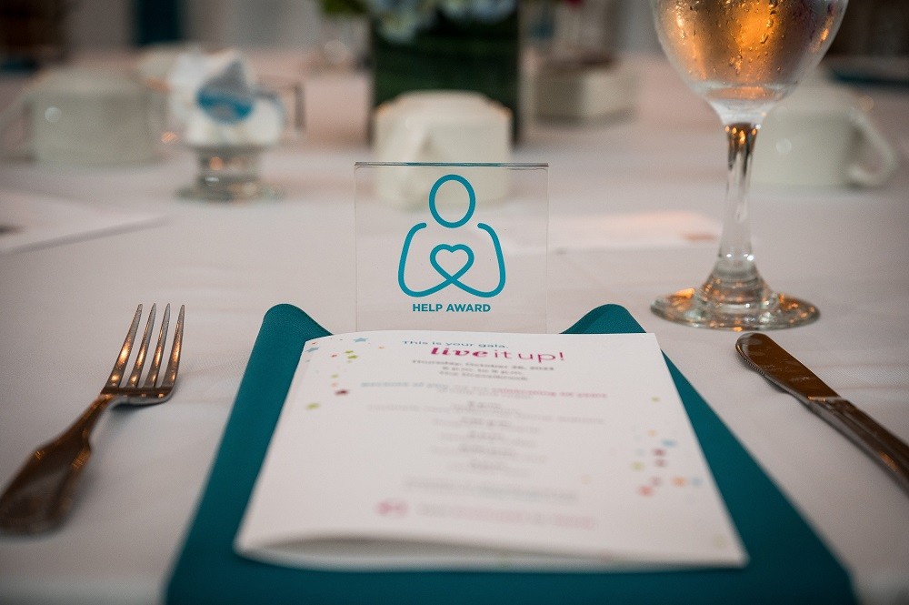 A photo of the table settings pre-dinner service at the 2023 Live It Up! gala. The single place setting features a white tablecloth, teal napkin, 2023 printed program booklet, knife and fork, water glass, and a small LIVE AWARD featuring the Help Hope Live logo pledge figure in teal.