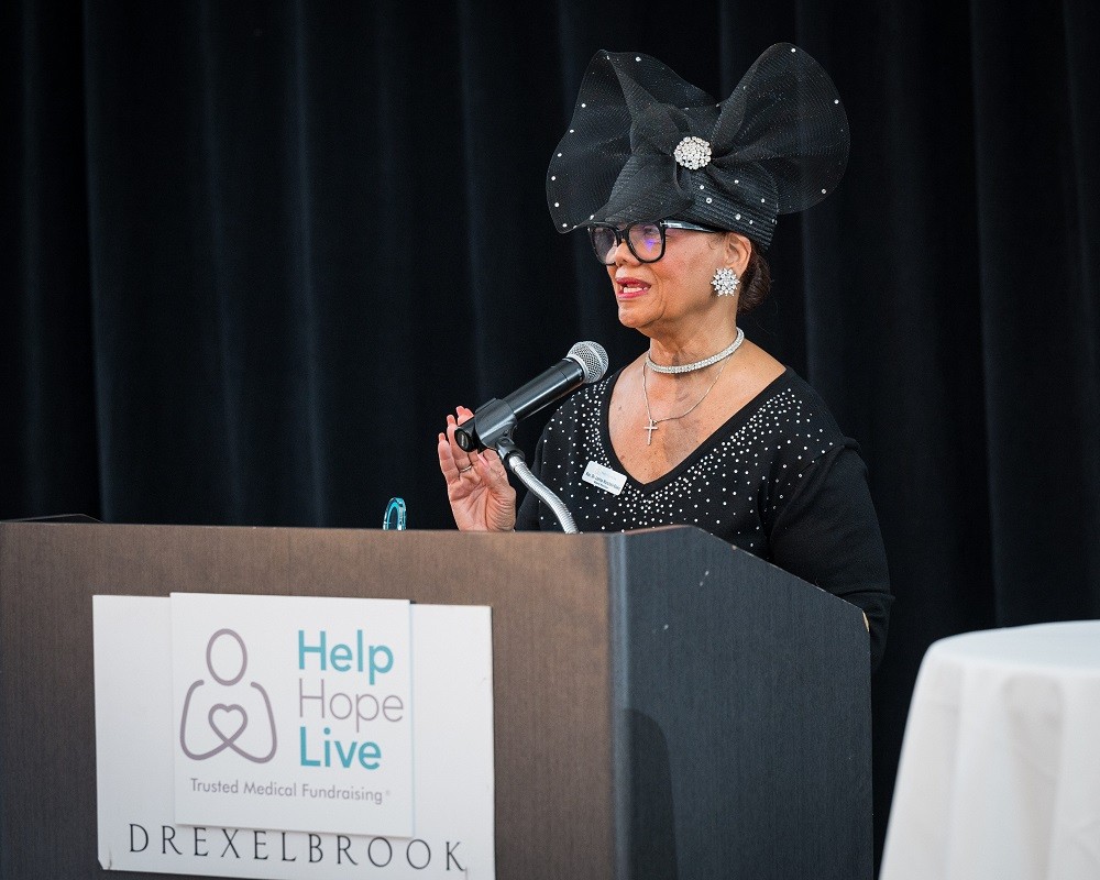 Rev. Dr. Lorina Marshall-Blake accepts her award at the podium at the 2023 Live It Up! event. Rev. Dr. Marshall-Blake has brown skin, glasses, a black long-sleeved dress, and a large eye-catching black hat with tulle detail.