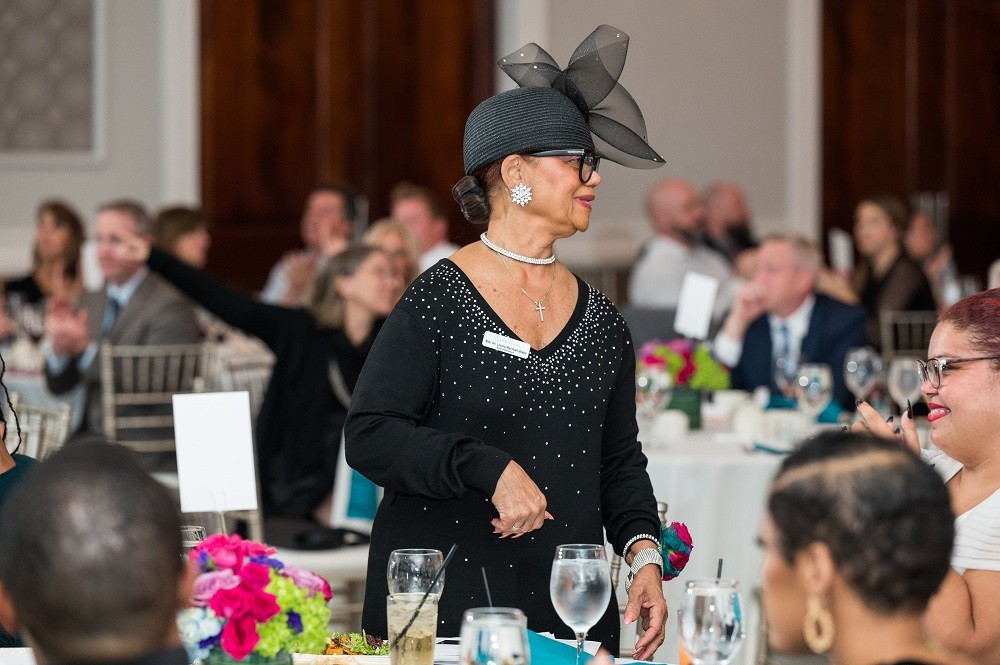 Tables of guests at the 2023 Live It Up! gala applaud as Rev. Dr. Lorina Marshall-Blake stands at her table. Rev. Dr. Marshall-Blake has brown skin, glasses, a black long-sleeved dress, and a large eye-catching black hat with tulle detail. The tables are set with crystalware, vibrant centerpiece flowers in magenta and light green, and white tablecloths.