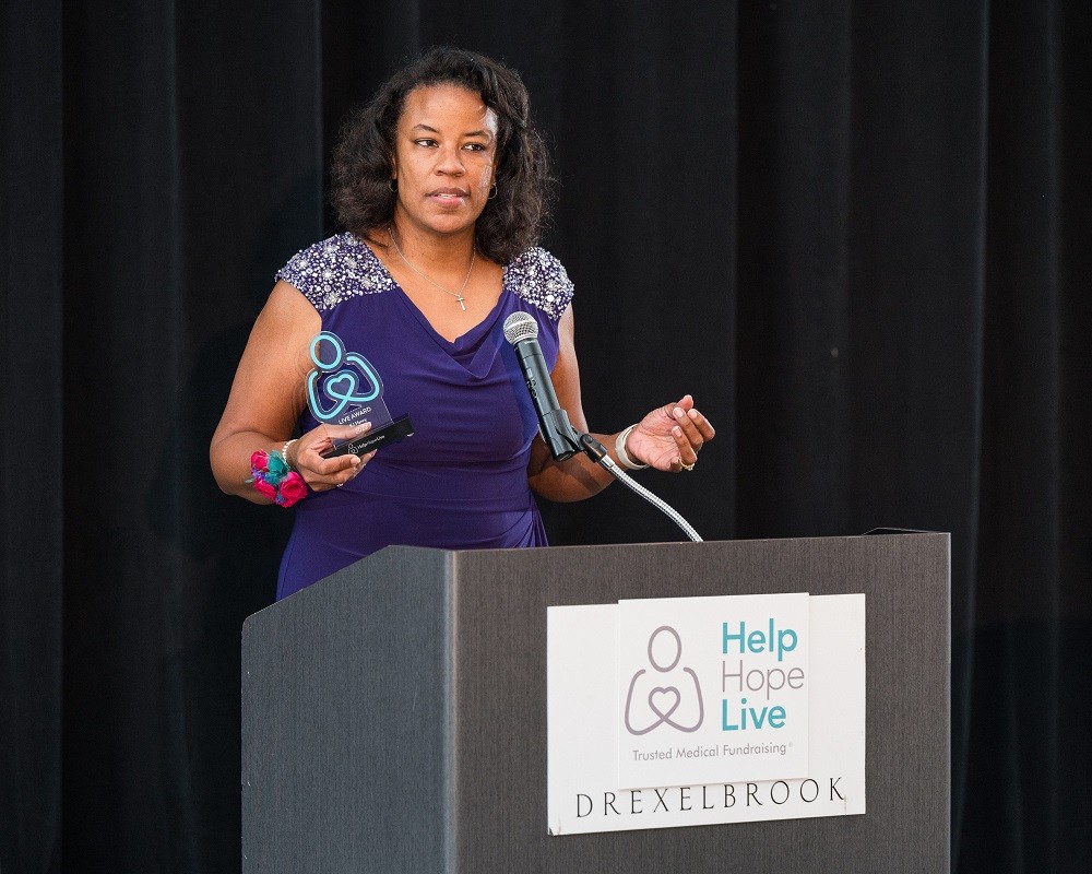 Nicole Henry is speaking at the podium at the 2023 Live It Up! gala with her award in her right hand (a teal Help Hope Live logo pledge figure). Nicole has brown skin, curly shoulder-length brown hair, and a purple cocktail dress.