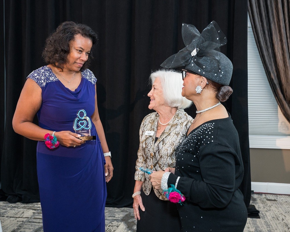 Award recipient Nicole Henry meets Help Hope Live co-founder Patricia Kolff and award recipient Rev. Dr. Lorina Marshall-Blake. Standing a head taller than Rev. Dr. Marshall-Blake, Nicole has brown skin, curly shoulder-length brown hair, and a purple cocktail dress. Rev. Dr. Lorina Marshall-Blake has brown skin, glasses, a black long-sleeved dress, and a large eye-catching black hat with tulle detail. Patricia Kolff has light skin, short white hair, black pants, and a formal gray white patterened blouse.