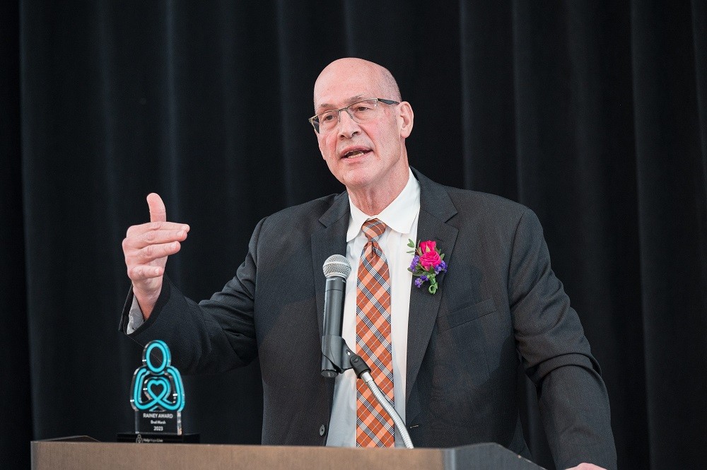 Brad Marsh is in mid-speech with his Live It Up! 2023 gala award on the podium in front of him. He has light skin, a bald head, glasses, and a black suit with an argyle tie.