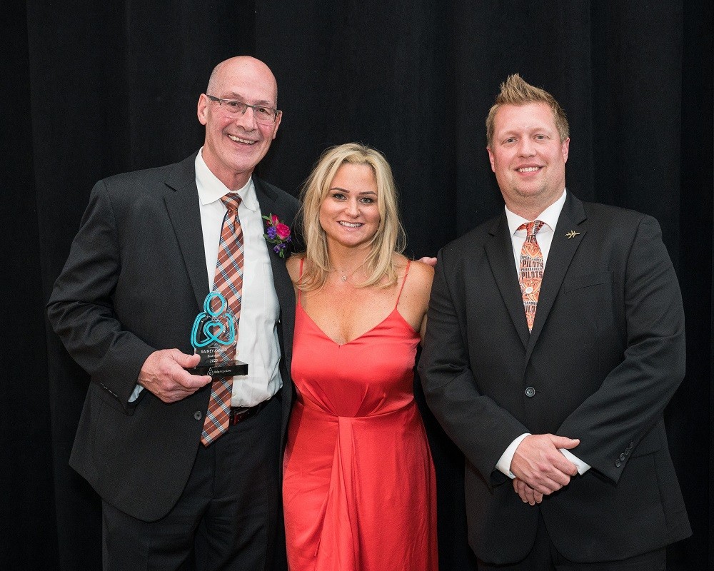 Volunteer of the Year Brad Marsh is with two other individuals as he holds his award at the 2023 Live It Up! gala. Brad Marsh has light skin, a bald head, glasses, and a black suit with an argyle tie. Next to him is a woman with light skin, blonde hair to her shoulders, and a red-orange sleeveless cocktail dress. Beside her is a man with light skin, short spiked light brown hair, and a black suit.