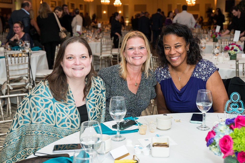 Three guests are seated at their table at the 2023 Live It Up! gala. The first guest is client Ambassador Linda Jara with light skin and a teal and tan vibrant geometric patterned formal shirt. To her left is sister Maggie with light skin, shoulder length blonde hair, and a blue dress. To Maggie's left is Nicole Henry, award recipient and client family member, who has brown skin, curly shoulder-length brown hair, and a purple cocktail dress.