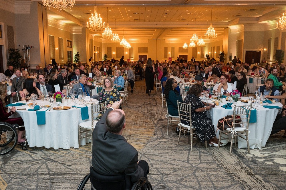 Mark Chilutti is seated in his wheelchair in a black suit visible from the back as he points into the crowd during the live Call to Fund portion of the 2023 Live It Up! gala. Before him are dozens of tables with white tablecloths and teal napkins in a carpeted ballroom with glowing chandeliers.