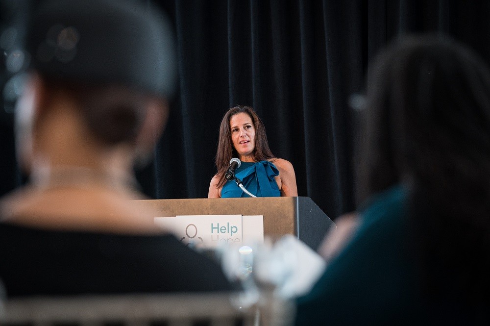 Help Hope Live Executive Director Kelly L Green is speaking at the podium at the 2023 Live It Up! gala. Kelly has light skin, a teal sleeveless halter ruffled cocktail dress, and dark staight hair past her shoulders. She is visible past the blurry partial outlines of two guests in the crowd listening.