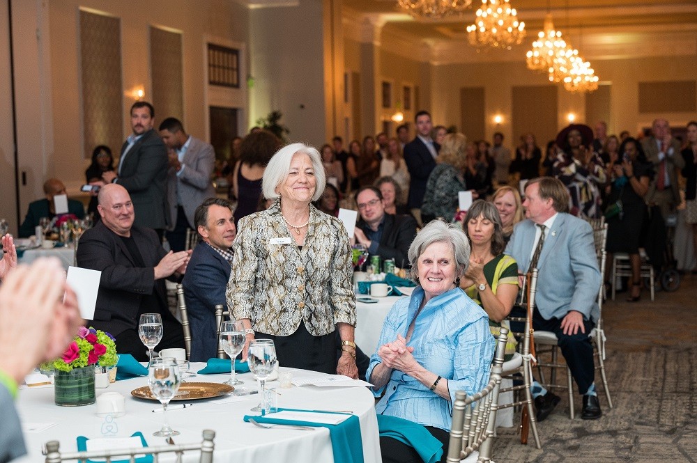 In a carpeted ballroom, the crowd of guests seated at white-tablecloth-covered tables at the 2023 Live It Up! gala are in the process of standing to give co-founder Patricia Kolff a standing ovation. Pat is standing at her table. She has light skin, white straight short hair, black pants, and a gray and white patterned formal blouse.