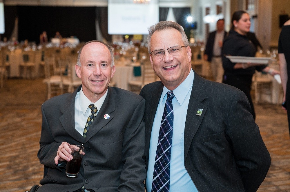 Mark Chilutti is seated in his wheelchair with Help Hope Live Ambassador Bill Soloway beside him at the 2023 Live It Up! gala. They are in a carpeted ballroom with tables and a projector screen visible behind them. Both men have black suits and navy ties, light skin, and short gray hair. Bill wears glasses. Mark holds a drink with a straw in a glass in his right hand.