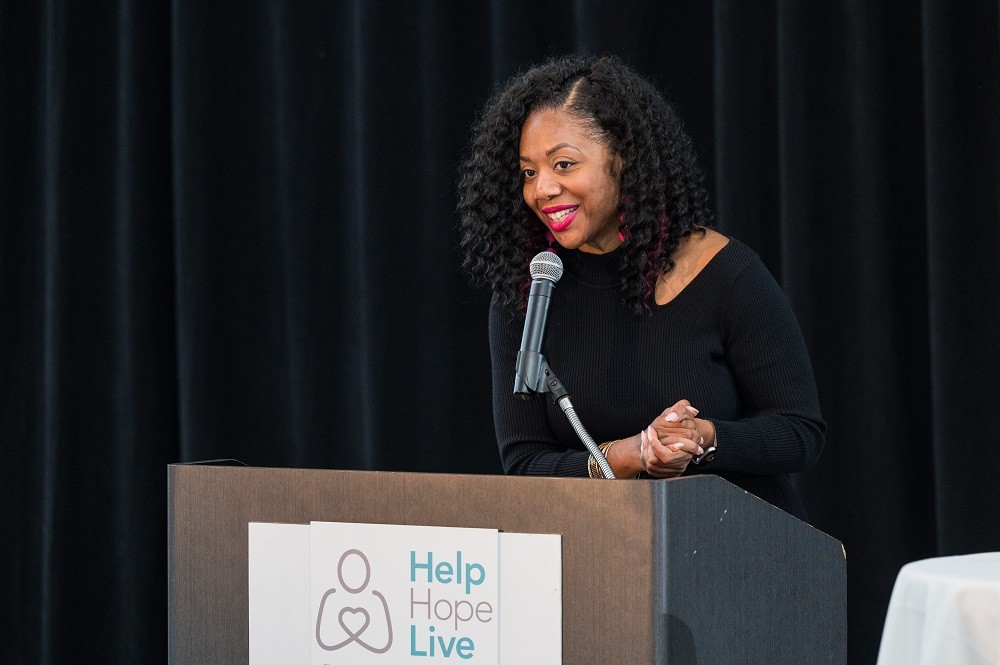 2023 Live It Up! gala emcee Cherri Gregg is speaking at the podium. She has brown skin, curly black hair to her shoulders, and a black long-sleeved formal outfit with red lipstick.