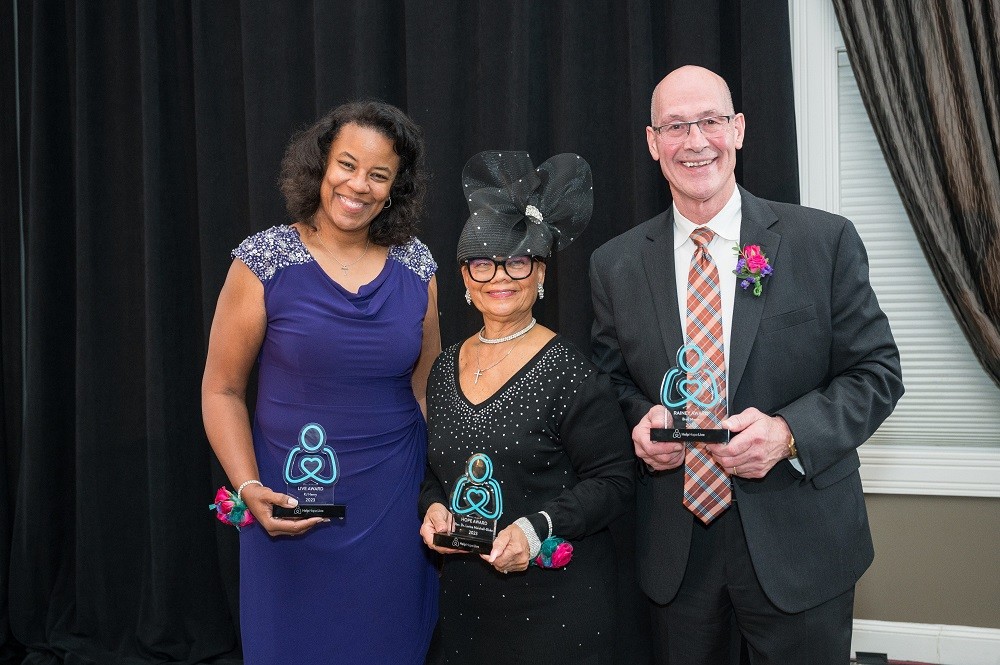 Our three 2023 Help Hope Live Awards honorees hold their awards, which are teal Help Hope Live logo pledge figures. The first is Nicole Henry who has brown skin, curly shoulder-length brown hair, and a purple cocktail dress. The second is Rev. Dr. Lorina Marshall-Blake, who has brown skin, glasses, a black long-sleeved dress, and a large eye-catching black hat with tulle detail. The third is Brad Marsh, who has light skin, a bald head, glasses, and a black suit with an argyle tie.