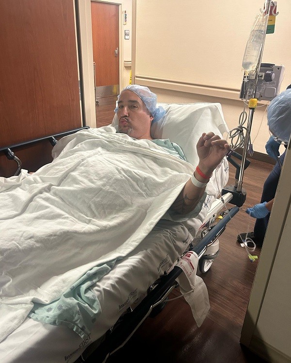 Kevin Lopez lies in a hospital bed hooked up to an IV cart with a light green hospital gown and a light blue transparent hair cap. He has hospital bracelets on his left wrist, light brown skin, a salt-and-pepper goatee, and dark eyes, and he's making a funny pursed-lip expression though his eyes look tired.