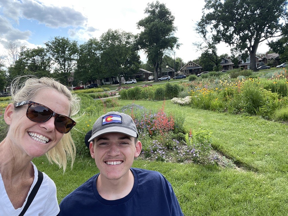 Spinal cord injury survivor Collin Bosse is seated in his wheelchair with his mom beside him taking a selfie. They are outdoors in a beautiful green space with wild flowers. Collin is a young man with light skin, a baseball cap, and a navy shirt. His mom has light skin, blonde dyed hair in a ponytail, sunglasses, and a white V-neck shirt.