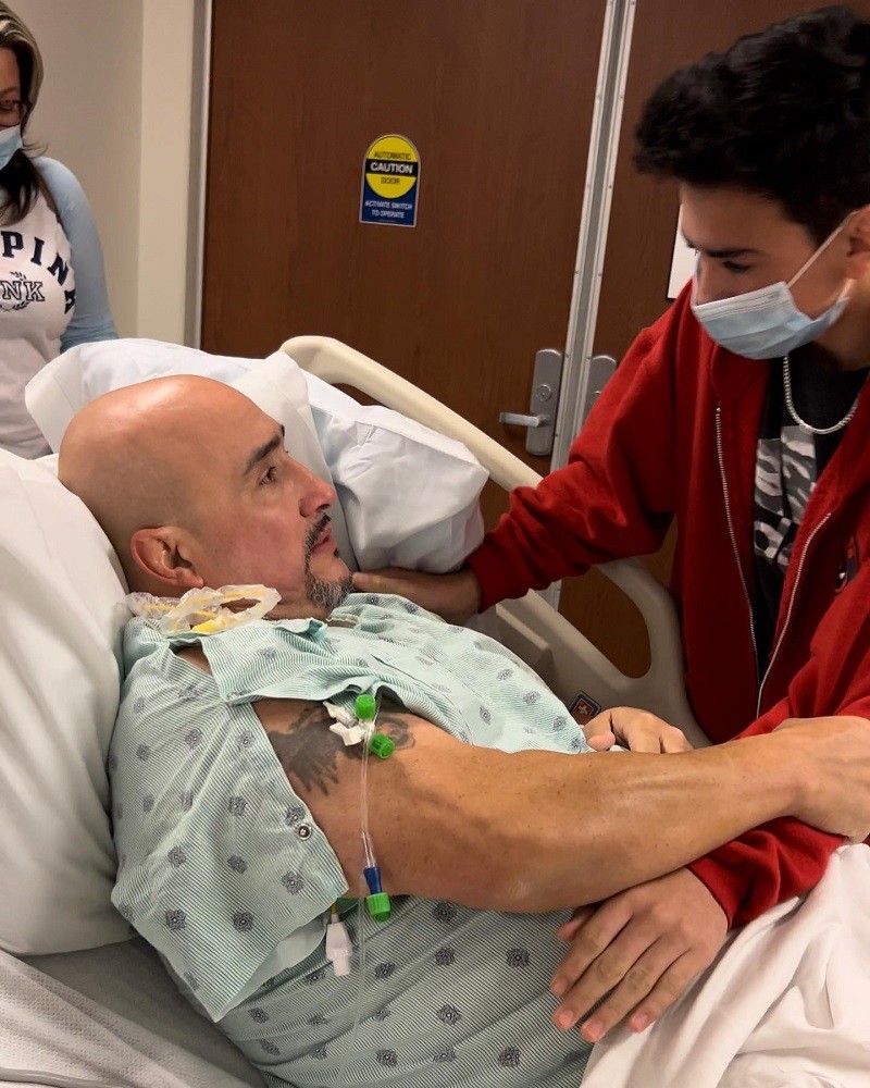 In an extremely emotional and intimate captured moment, Kevin Lopez lies in a hospital bed and clutches his son's arm in his right hand, looking into his son's eyes with an expression of intense love. Kevin has light brown skin, a bald head, dark eyes, and a salt-and-pepper goatee. His son has light brown skin, dark eyes, short dark hair, a blue medical face mask, and a red hoodie.