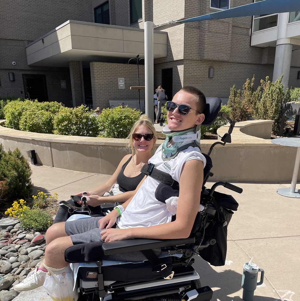 Collin Bosse is seated outside a facility in his black power chair with a strap across his chest and a neck brace. Collin is a young man with light skin, sunglasses, and short cropped dark hair. He is with his sister, who has light skin, a black tank top, sunglasses, and blonde hair in a ponytail.