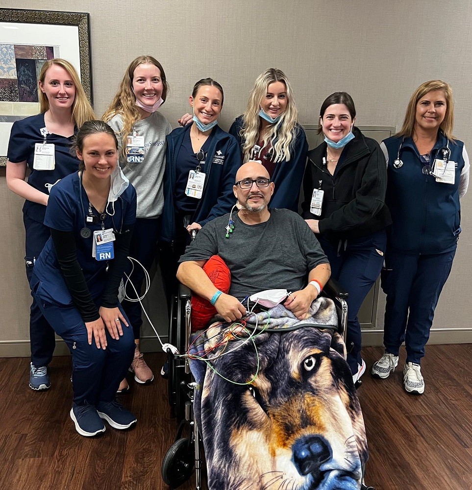 Kevin Lopez is seated in a wheelchair connected to medical equipment with a wolf face blanket over his lap, surrounded by seven female RN medical staff members, all wearing navy scrubs. Kevin has light brown skin, a bald head, dark eyes, black rimmed glasses, and a salt-and-pepper goatee. He and all the female RNs around him are smiling.