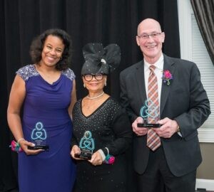 Our three 2023 Help Hope Live Awards honorees hold their awards, which are teal Help Hope Live logo pledge figures. The first is Nicole Henry who has brown skin, curly shoulder-length brown hair, and a purple cocktail dress. The second is Rev. Dr. Lorina Marshall-Blake, who has brown skin, glasses, a black long-sleeved dress, and a large eye-catching black hat with tulle detail. The third is Brad Marsh, who has light skin, a bald head, glasses, and a black suit with an argyle tie.