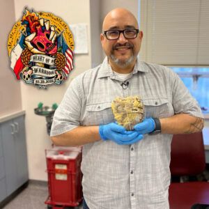 Kevin Lopez is smiling as he literally holds his old heart in his hands, raising it to the level of his own heart in blue medical gloves. The heart is yellow-gray and cut into slices but kept together with a band, and it is larger than an orange. He appears to be in a hospital check-up style room. He has light brown skin, a bald head, dark eyes, a salt-and-pepper goatee, and black-rimmed glasses, and he wears a gray and white striped button-up short-sleeved shirt. Over the photo in the top-left corner is a logo of a hand holding a sugar skull detailed heart with a fuse like a grenade, all in shocking pink with yellow fine detail. The hand is held in a skeleton's grasp with gold marigolds, additional sugar skulls, the Texas and American flags, and the text Heart of a Warrior 10.23.2022.