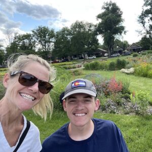 Spinal cord injury survivor Collin Bosse is seated in his wheelchair with his mom beside him taking a selfie. They are outdoors in a beautiful green space with wild flowers. Collin is a young man with light skin, a baseball cap, and a navy shirt. His mom has light skin, blonde dyed hair in a ponytail, sunglasses, and a white V-neck shirt.