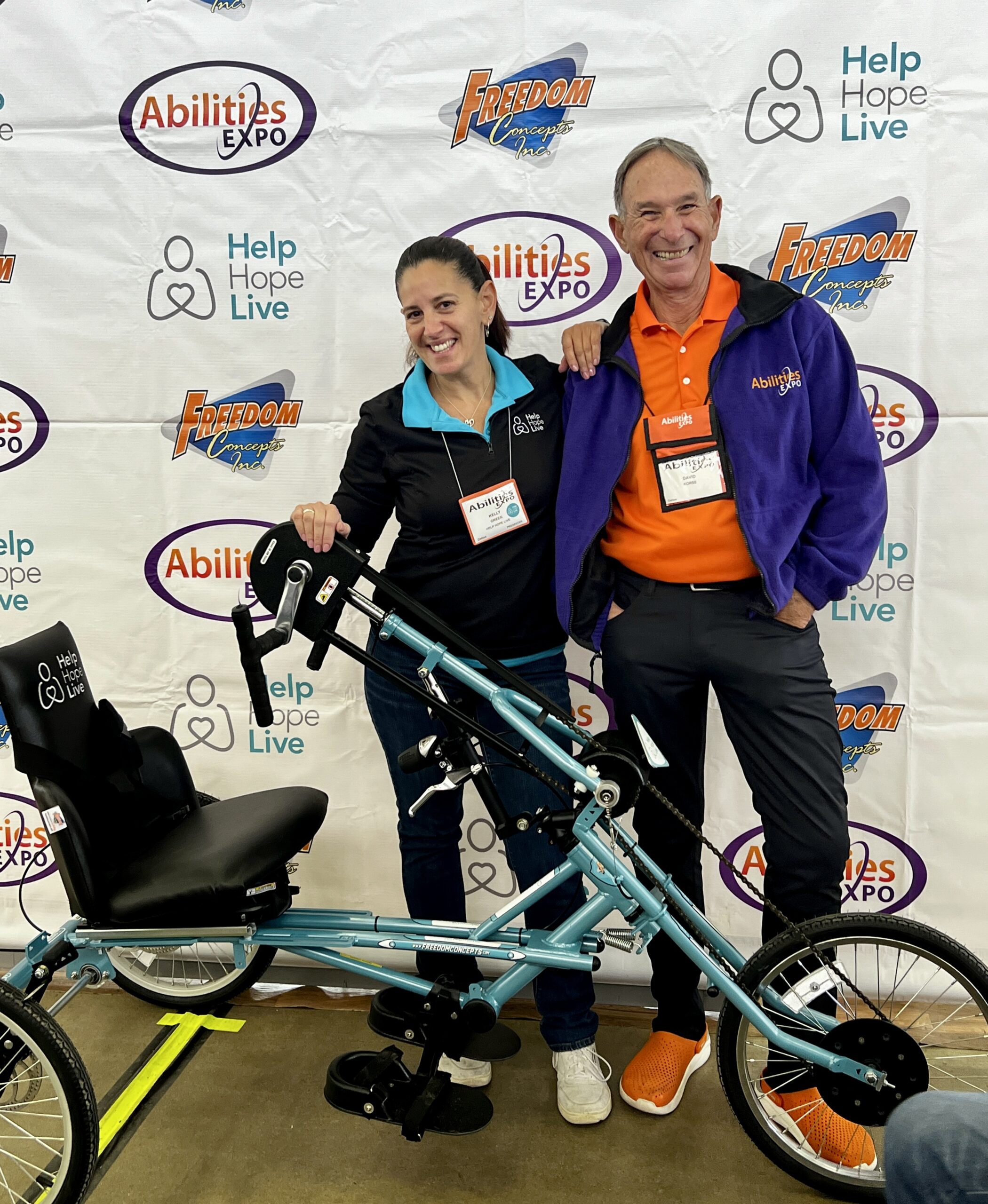 Kelly L Green stands with an Abilities Expo employee in front of a step-and-repeat banner at Abilities Expo Dallas. In front of them is a teal, Help Hope Live-branded adaptive handcycle bike. Kelly has light skin and dark hair in a ponytail with a black Help Hope Live jacket and teal polo collar. The Abilities Expo employee has light skin, short gray hair, a vibrant orange polo and sneakers, and a purple Abilities Expo jacket.