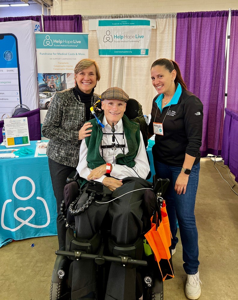 Kelly L Green stands next to client Stephen Mackintosh and a loved one at Abilities Expo Dallas in front of the teal Help Hope Live booth and banners. Kelly has light skin and dark hair in a ponytail with a black Help Hope Live jacket and teal polo collar. Stephen has light skin and short white hair with a tartan cap and white collared shirt. He is seated in his black power chair. His loved one has light skin and short golden blonde hair.