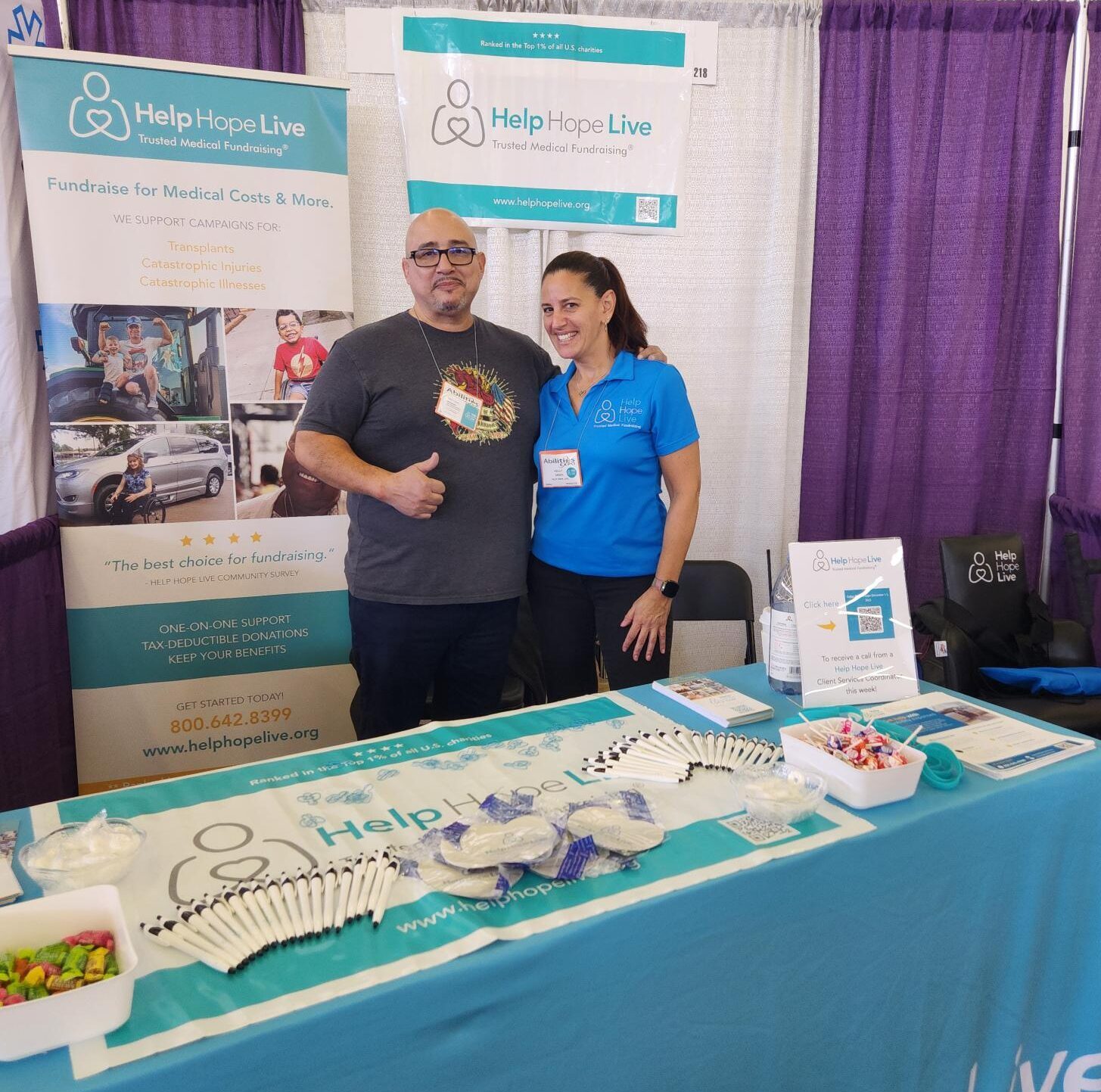 Kevin Lopez and Kelly L Green are at the Help Hope Live booth at Abilities Expo Dallas. There is a standing banner behind them featuring four photos of Help Hope Live clients and information about the nonprofit. On the table is pens, candy, jar openers in heart shapes with the Help Hope Live logo, and materials. Kevin has light brown skin, a gray goatee, a bald head, black rimmed glasses, and a gray Heart of a Warrior fundraising shirt with a stylized Dia de los Muertos or calavera style design. Kelly L Green has light skin and dark hair in a ponytail with a blue Help Hope Live logo polo shirt.