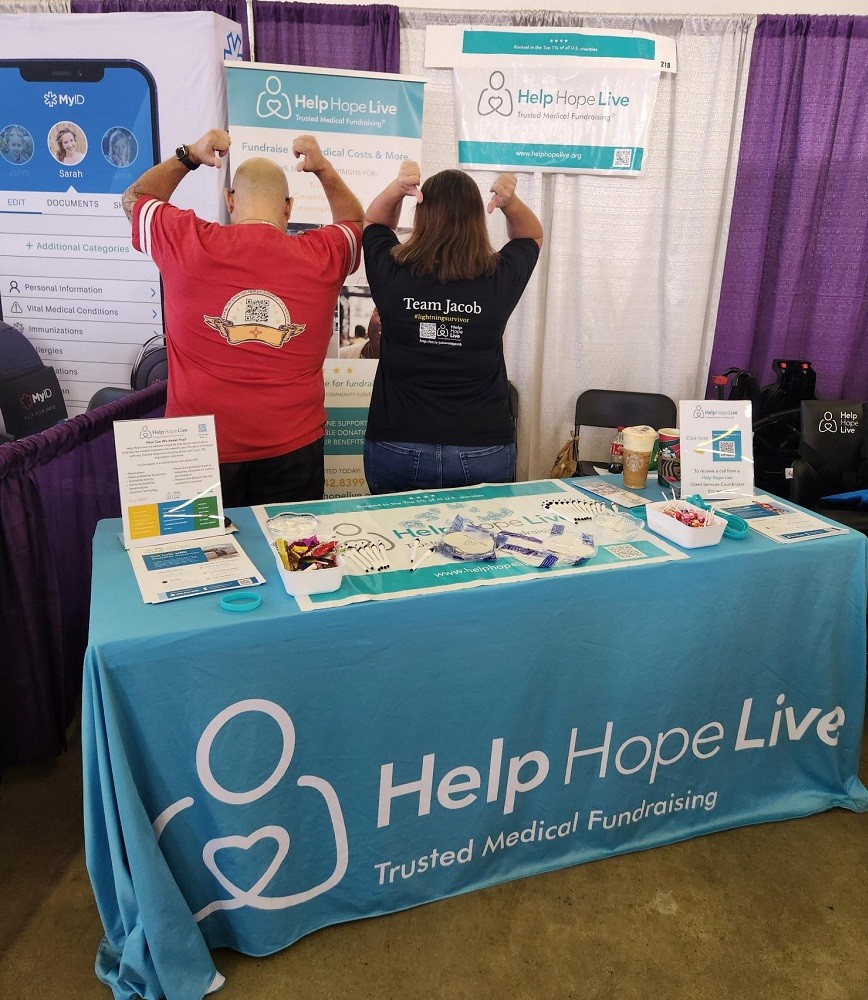 Kevin Lopez and Barbara Brewer, both Help Hope Live clients, are at the Help Hope Live booth with the teal tablecloth and logo visible in front of them. They are both facing backward and using their thumbs to point to the backs of their t-shirts. Kevin has light brown skin, a bald head, and a bright red t-shirt with a QR code on it (too far away to scan from this photo). Barbara has light skin, shoulder-length brown hair, jeans, and a black shirt that reads Team Jacob #lightningsurvivor. It also features a QR code, not scannable from this photo.