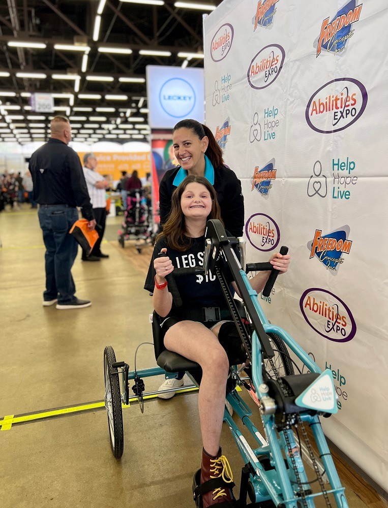 On the expo floor at Abilities Expo Dallas, Help Hope Live’s Kelly L Green is with 15-year-old Hallie, an adaptive bike recipient. Kelly has light skin, dark eyes, and dark hair in a ponytail. Hallie has light skin, a big smile with braces, and wavy shoulder length brown hair. She is operating her new adaptive bike with her hands—it is teal with Help Hope Live branding on the front.