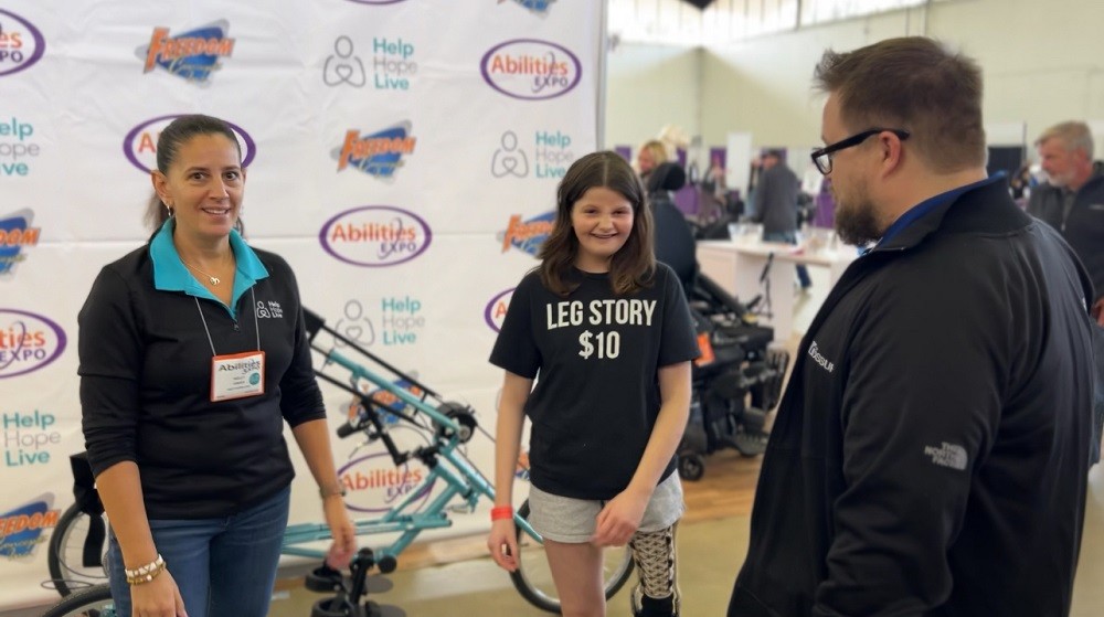 Kelly L Green and 15-year-old adaptive bike recipient Hallie are at Abilities Expo Dallas with a step-and-repeat banner behind them bearing logos for Freedom Concepts, Help Hope Live, and Abilities Expo. Hallie stands in front of her teal Help Hope Live themed handcycle adaptive bike. Hallie has light skin, a big smile with braces, and wavy shoulder length brown hair. Her left leg is a prosthetic leg, and she wears a black t-shirt that reads LEG STORY $10. Kelly has light skin and dark hair in a ponytail with a black Help Hope Live jacket and teal polo collar, and she’s making a funny “what the heck?” expression at the camera as Hallie laughs next to her.