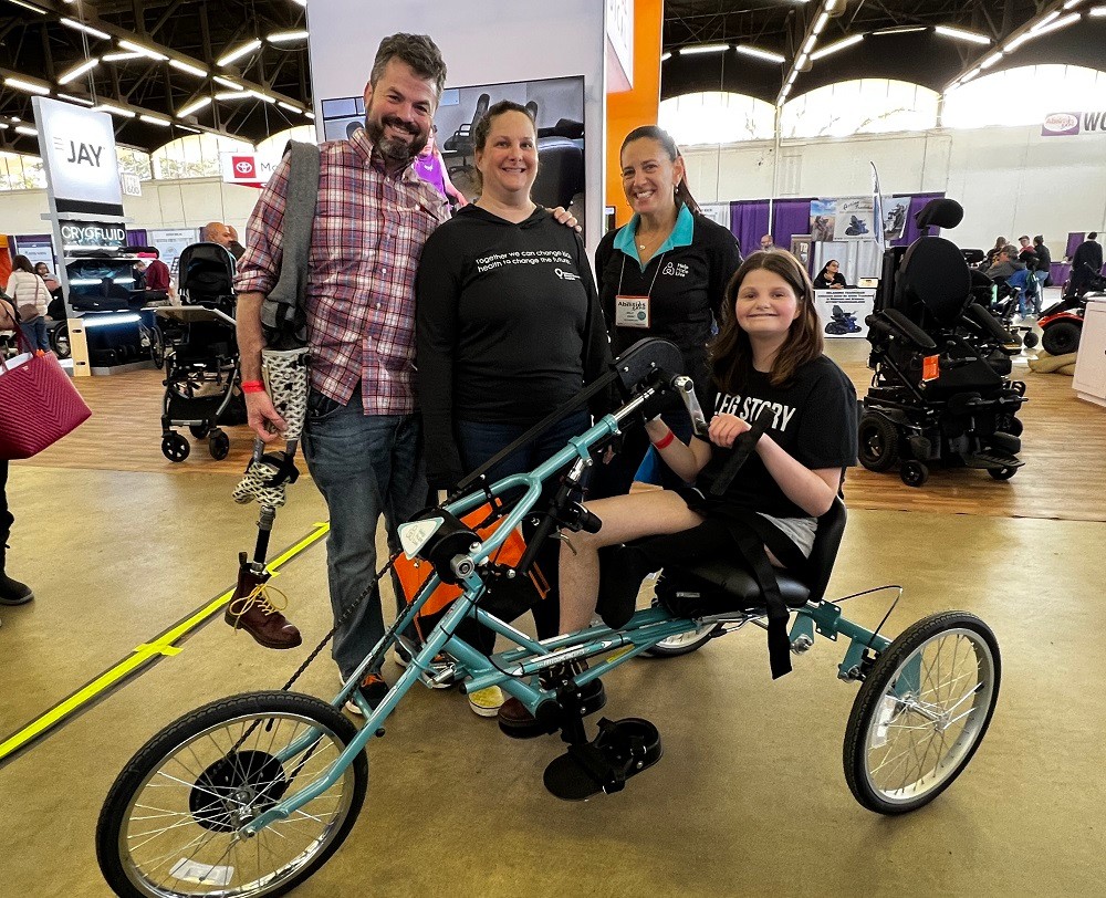 Kelly L Green is with 15-year-old Hallie and her parents at Abilities Expo Dallas. Hallie is sitting on her new teal Help Hope Live-themed handcycle bike. She has light skin, shoulder-length brown hair, a black t-shirt, and one leg. Her dad has light skin, thick gray hair, and a gray short beard. He wears jeans and a button-up shirt and carries Hallie’s prosthetic leg, which ends in a hiking boot. Her mom has light skin, brown hair pulled back, and a black athletic long-sleeved shirt. Kelly has light skin and dark hair in a ponytail with a black Help Hope Live jacket and teal polo collar.