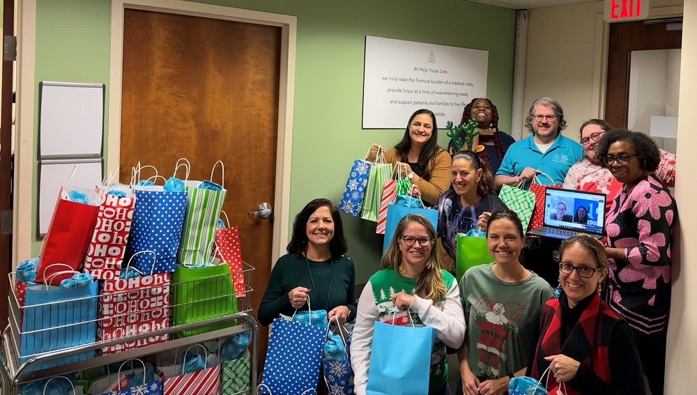 A photo of the Help Hope Live team together at the offices for the 2023 Bags of Hope packing. They are next to a cart of brightly colored full holiday gift bags and are holding additional bags in the photo. The 10 staff members are diverse in skin color and age with one male and nine females, plus 2 additional staff members on a laptop one of the in-person staff is holding up for the photo. Many of them are dressed in silly holiday sweaters or attire.
