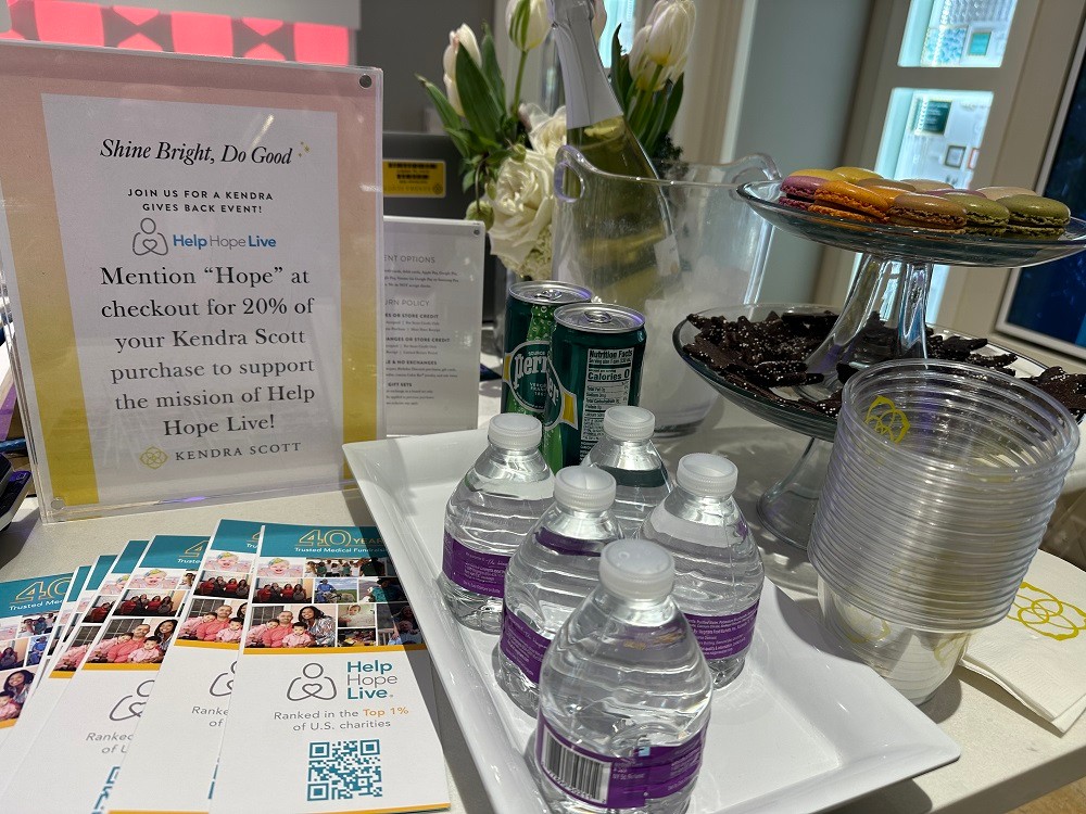 A Help Hope Live display table and snack assortment at a Kendra Scott fundraising event with brochures, water bottles and Perrier, peppermint bark, multi-colored macarons, and a sign reading Shine Bright, Do Good: Join Us for a Kendra Gives Back Event! Mention 