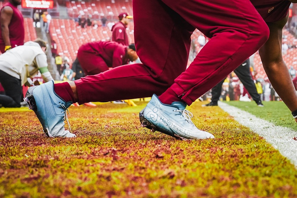 A photo of Commanders player KJ Henry's legs in red workout gear on the field wearing his Help Hope Live branded cleats. The cleats read Help Hope Live on the side with teal, white, and gray ombre detail.