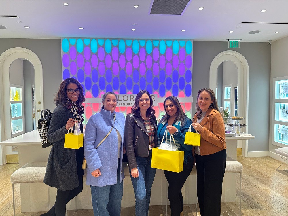 At a Kendra Scott store, five Help Hope Live staff and board members hold up their bright yellow Kendra Scott shopping bags. They are all women of various ages and ethnicities in business-casual winter wear. The shop looks modern and high-end with light wood floors, glass cases, and plush cream furniture.