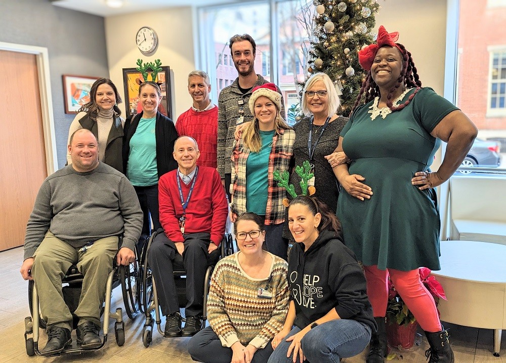 Members of the Help Hope Live staff and board with staff at Magee Rehabilitation in the Magee Rehabilitation facility with windows showing a city street and red brick buildings and a decorated holiday tree with silver ornaments. The individuals pictured are a diverse mix of ages and genders with most standing and two individuals seated in their wheelchairs. Many wear red, green, or other festive holiday touches.