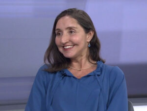 Heart transplant recipient Manoela Vieira is on a TV interview. She is wearing a blue blouse, earrings, and a necklace and she has light brown skin, red lipstick, and shoulder-length brown hair.