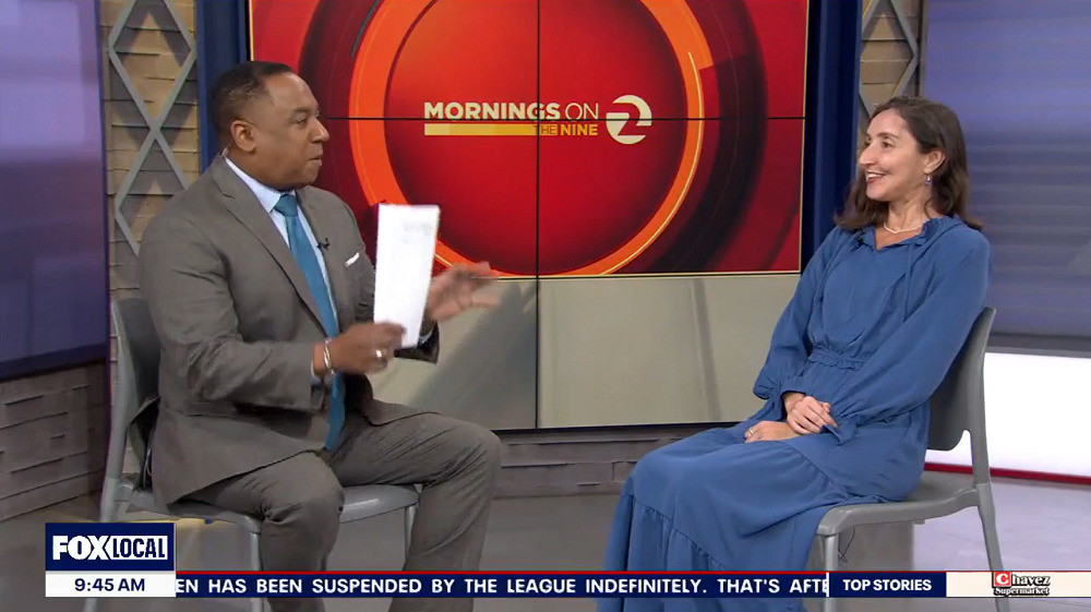 Manoela Vieira is interviewed on TV following a heart transplant. She and the program host are seated in gray chairs on a stage with a red TV screen behind them that reads Mornings on the Nine. A ticker along the bottom of the screen reads FOX Local 9:45 am. The host has brown skin, short black hair, and a gray suit with a blue tie and holds paper in his right hand. Manoela has light brown skin, shoulder-length brown hair, red lipstick, brown eyes, and a blue flowing dress with long sleeves.