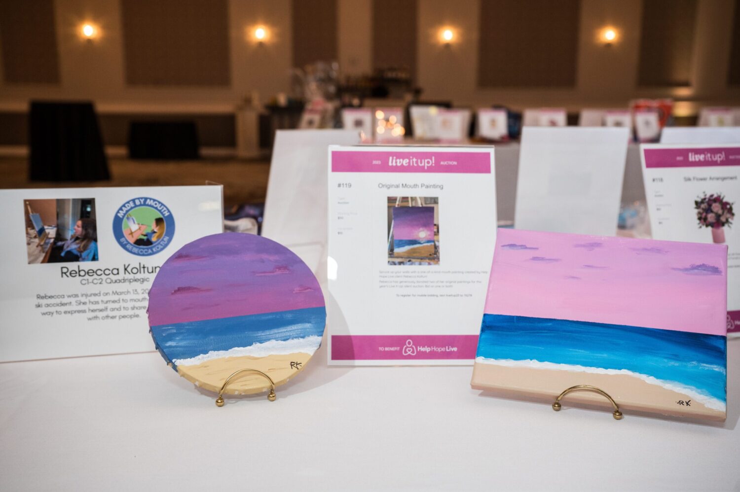 Two mouth-painted artworks by Rebecca Koltun at the auction table for Live It Up! 2023. They are beach scenes at sunset or sunrise with a pink-purple sky and bright blue water next to brown sand.