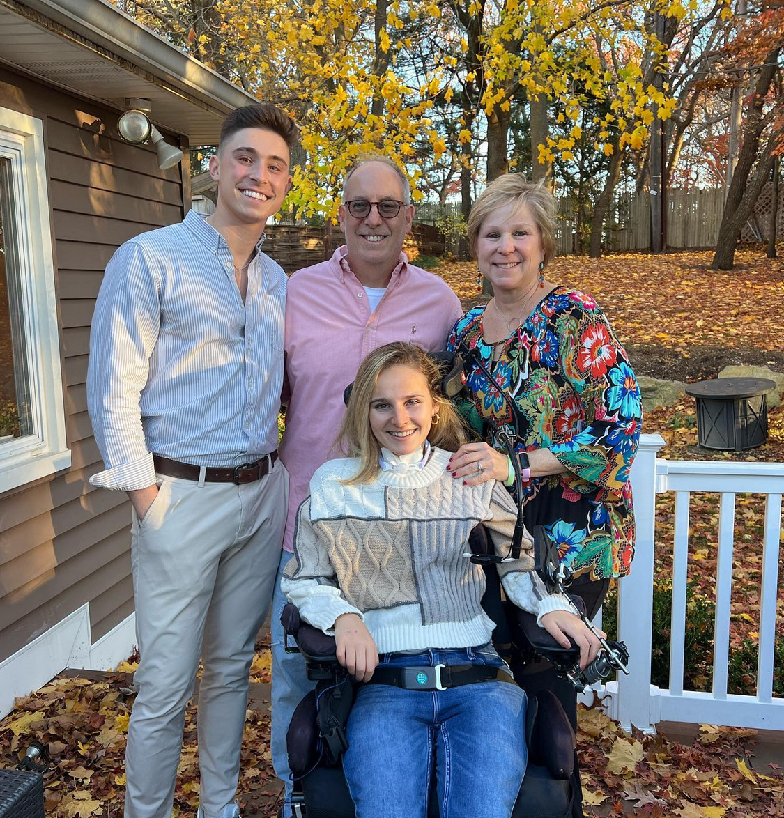 Rebecca with her family outdoors on a deck with fall leaves visible in the yard behind them. Seated in her black power chair, Rebecca has light skin, shoulder-length brown and blonde hair, a turtleneck graphic print tan and cream sweater, and jeans. Behind her stand her brother with light skin, a collared gray shirt, khaki pants and a belt with light skin and short brown hair. Her father, with short gray hair, sunglasses, and a pink button-up shirt. Her mom, with light skin, short sandy brown hair, and a vivid graphic print abstract flower long-sleeved shirt.