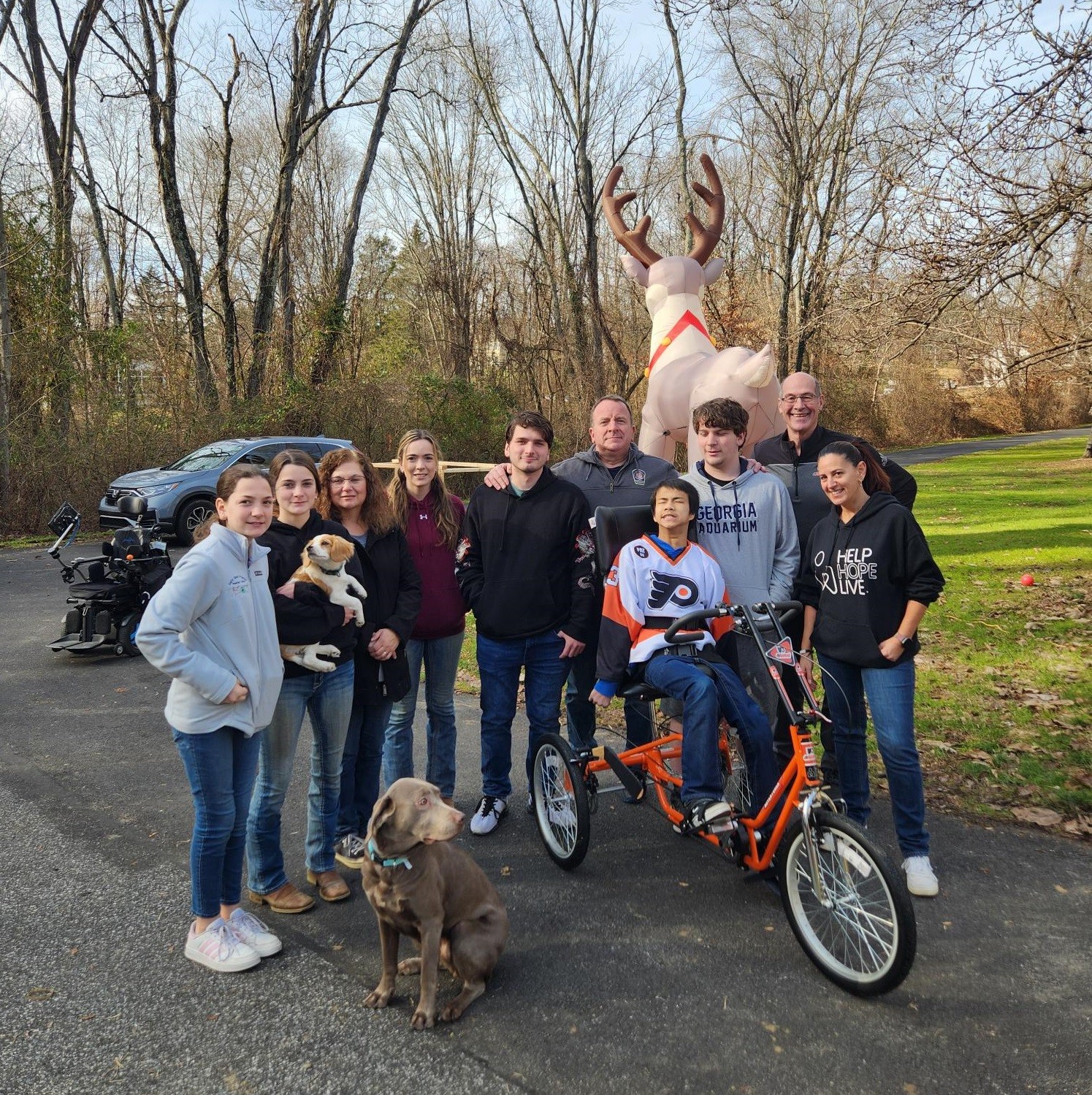 Teenager Joshua Johnson smiles and squints as he sits on his orange three-wheel adaptive bike. He is Asian with dark hair and a Philadelphia Flyers orange, black, and white jersey. He is surrounded by Kelly Green of Help Hope Live, Brad Marsh of the Flyers Alumni, and friends and family of various ages plus a gray-brown dog. They are in a driveway with a car, a giant inflatable reindeer, a lawn, and a black empty power chair visible behind them.