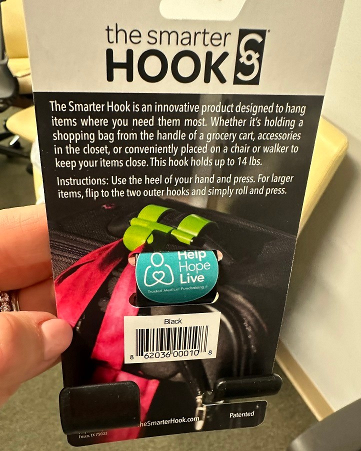 The back of the packaging for a The Smarter Hook. It is a black ring with the Help Hope Live logo and tagline Trusted Medical Fundraising over it. It reads The Smarter Hook is an innovative product designed to hang items where you need them most. Whether it's holding a shopping bag from the handle of a grocery cart, accessories in the closet, or conveniently placed on a chair or walker to keep your items close. This hook holds up to 14 pounds. Instructions: Use the heel of your hand and press. For larger items, flip to the two outer hooks and simply roll and press.