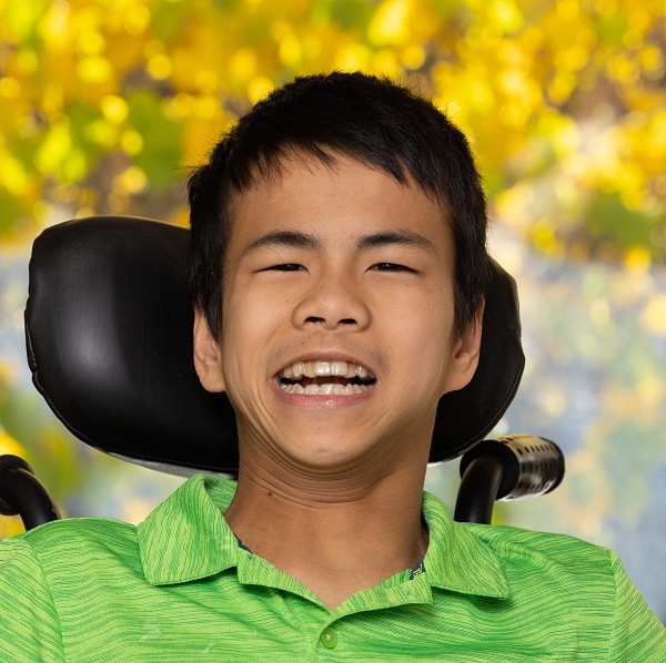 In a portrait, teenager Joshua Johnson is Asian with dark eyes, black short hair, and a big toothy smile as he sits in a black power chair.