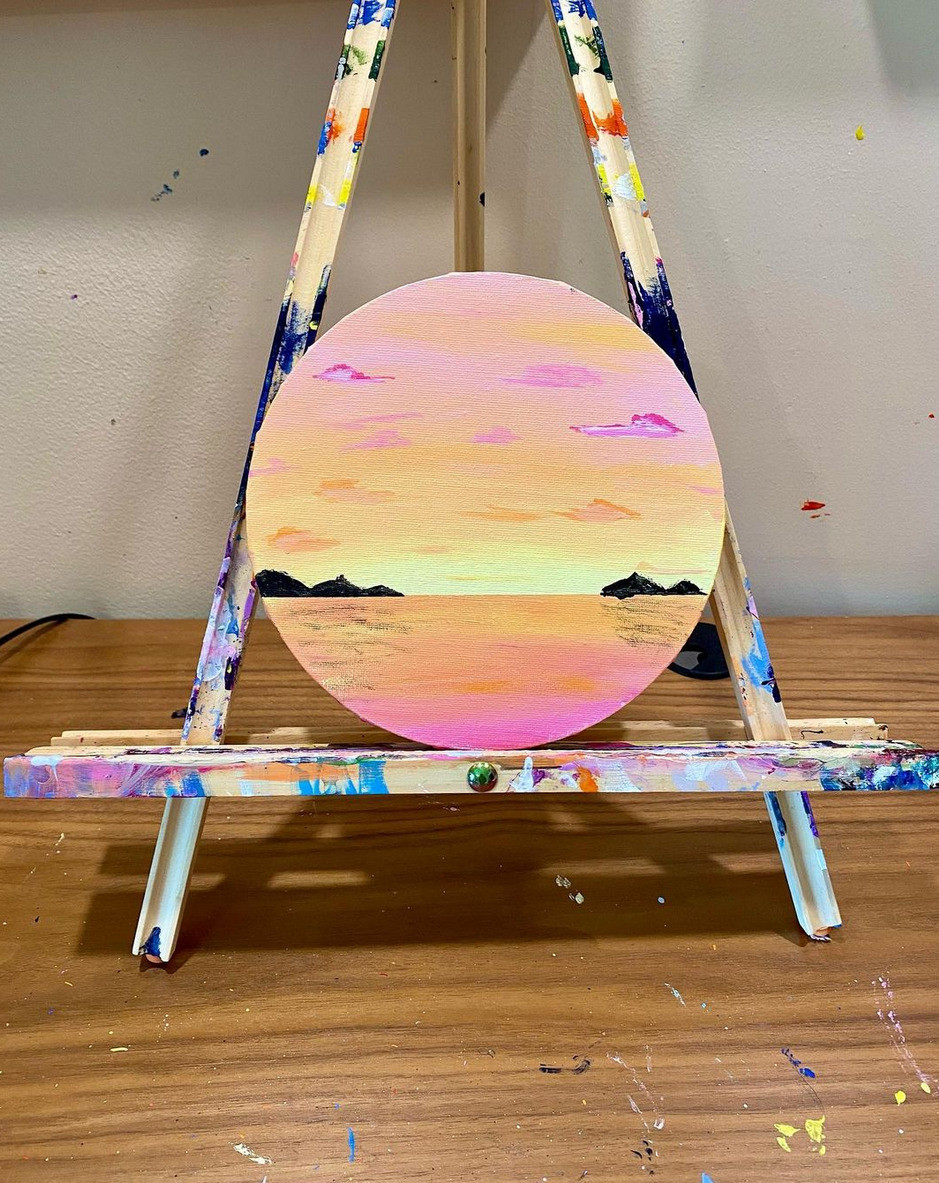 A Rebecca Koltun mouth-painted piece, circular on a small easel covered in paint splatters. The piece is a sunrise or sunset over water featuring calming shades of pink, orange, and yellow.